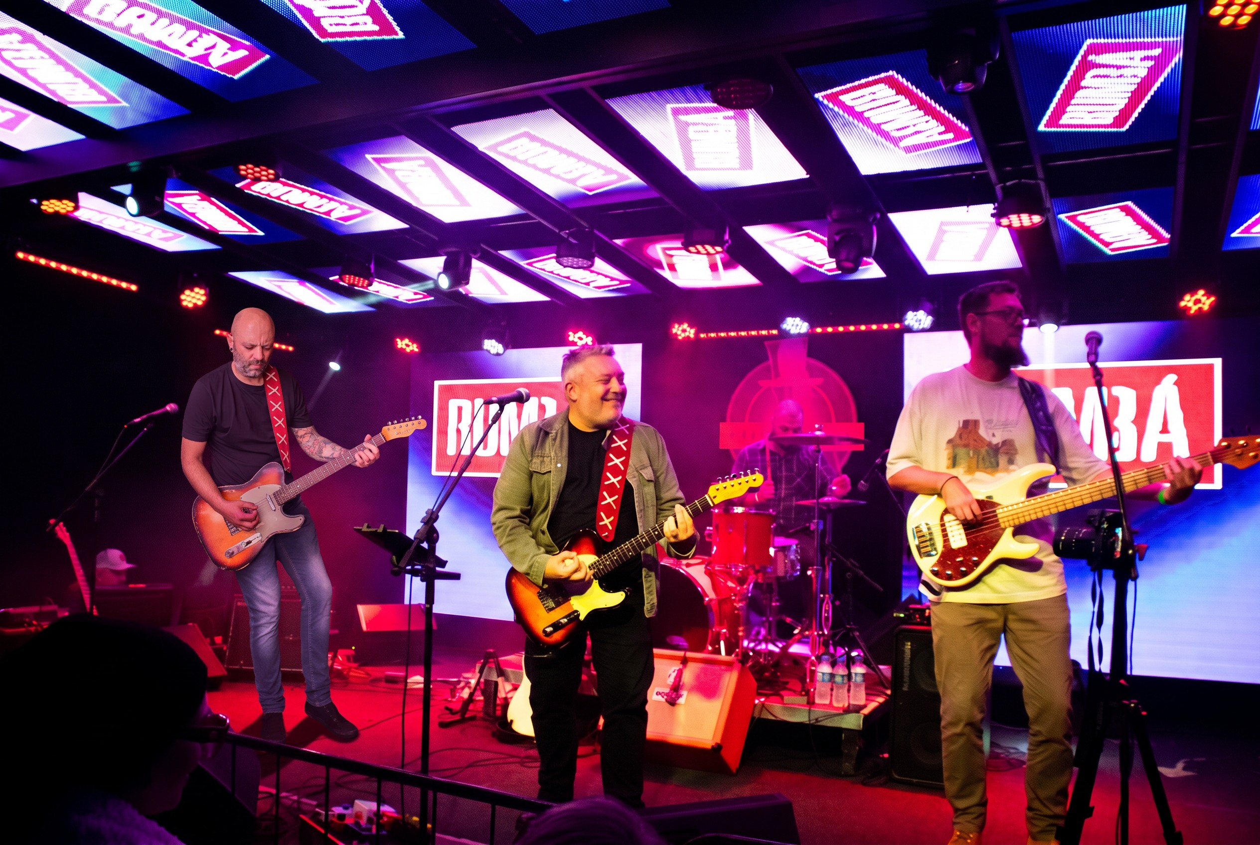 Os quatro músicos da Rumbá ao vivo no palco, com letreiros RUMBÁ no fundo